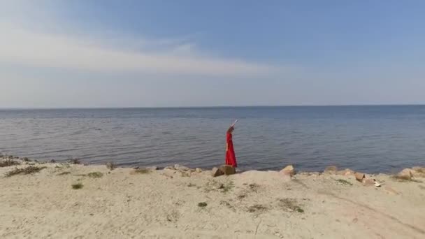 Graseful linda mujer joven en rojo vestido largo bailando levanta las manos sobre la piedra grande cerca de la vista espectacular de agua de mar azul y bosque increíble. Disparando con dron. Movimiento lento . — Vídeos de Stock