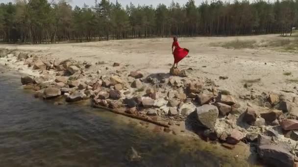 Ніжна вдягнена жінка в червоне довге плаття піднімає руки на великий камінь біля ефектного вигляду блакитної морської води і дивовижного неба. Сучасна танцівниця практикує на відкритому повітрі. Стрілянина на дроні — стокове відео