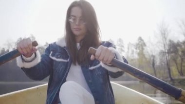 Beyaz pantolon, kot ceket ve güneş gözlüğü nehir üzerinde sarı tekne üzerinde kürekler Pretty glamour kadın, etrafa bakıyor. Kürek çekmeyi öğrenen kız. Doğayla bağlantı. Aktif yaşam tarzı