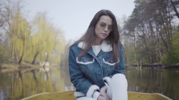 Portrait d'une magnifique jeune femme en lunettes de soleil et une veste en jean flottant sur un bateau sur un lac ou une rivière. Belle brune se relaxe activement un jour de congé ou de voyage en profitant de la nature . — Video