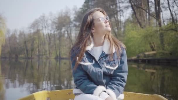 선글라스를 착용한 우아한 젊은 여성의 초상화와 호수나 강위에 떠 있는 데님 재킷. 아름다운 갈색 머리는 적극적으로 하루 쉬거나 자연을 즐기는 여행에서 휴식을 취하고 있습니다.. — 비디오