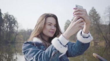 Nehirde teknede oturan ve cep telefonuyla selfie çeken güzel genç kadının portresi. Arka planda güzel manzara. Doğayla bağlantı. Aktif yaşam tarzı