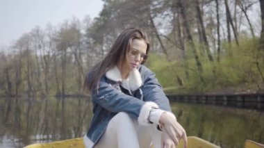 Nehirde teknede beyaz pantolon ve kot ceket ile Oldukça çekici kadın, güneşin tadını çıkarıyor. Arkasında güzel manzara. Doğayla bağlantı. Aktif yaşam tarzı. Bir kız teknede yüzüyor..