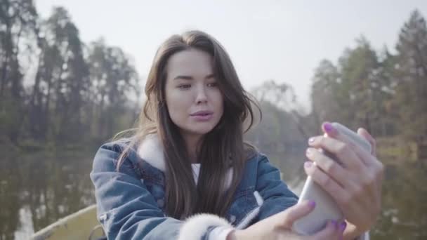 Ritratto di donna piuttosto glamour seduta sulla barca sul fiume a farsi un selfie sul cellulare. Bellissimo paesaggio sullo sfondo. Connessione con la natura. Stile di vita attivo — Video Stock