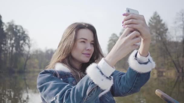 Το πορτρέτο της ομορφης νεαρής γυναίκας που κάθεται στη βάρκα στο ποτάμι και παίρνει selfie στο κινητό της. Όμορφο τοπίο στο παρασκήνιο. Σύνδεση με τη φύση. Ενεργός τρόπος ζωής — Αρχείο Βίντεο