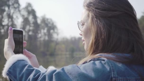 Вид ззаду красива гламурна жінка в джинсовій куртці і окулярах, фотографуючи красиву природу на своєму мобільному телефоні. Прекрасний краєвид навколо. Зв'язок з природою — стокове відео