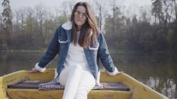 Retrato de uma mulher bonita encantadora em óculos e uma jaqueta de ganga flutuando em um barco em um lago ou rio. Linda morena está relaxando ativamente em um dia de folga ou viajando apreciando a natureza . — Vídeo de Stock