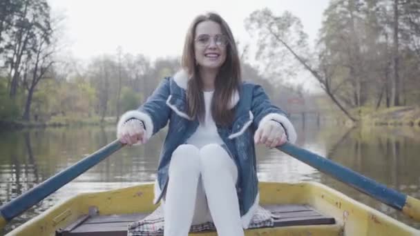 Hübsche junge Glamour-Frau in weißer Hose, Jeansjacke und Sonnenbrille paddelt auf dem gelben Boot auf dem Fluss und schaut sich um. Mädchen lernen Rudern. Verbindung mit der Natur. Aktiver Lebensstil — Stockvideo