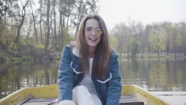 Retrato de una encantadora mujer linda en gafas y una chaqueta de mezclilla flotando en un barco en un lago o río. Hermosa morena es activamente relajarse en un día libre o viajar disfrutando de la naturaleza . — Vídeos de Stock