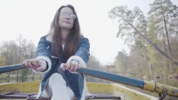 Porträt einer charmanten jungen Frau mit Brille und Jeansjacke, die auf einem Boot auf einem See oder Fluss treibt. schöne Brünette ist aktiv entspannen an einem freien Tag oder Reisen genießen die Natur. — Stockvideo