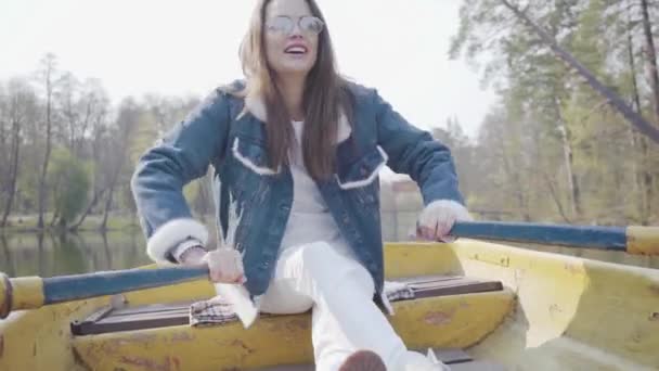 Die hübsche lächelnde Glamour-Frau in weißer Hose, Jeansjacke und Sonnenbrille paddelt mit überkreuzten Beinen auf dem gelben Boot auf dem Fluss. Mädchen lernen Rudern. Verbindung mit der Natur. aktiv — Stockvideo