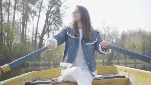 Portrait of a cute girl in glasses and a denim jacket floating on a boat on a lake or river. Beautiful brunette is actively relaxing on a day off or traveling enjoying nature. — Stock Video