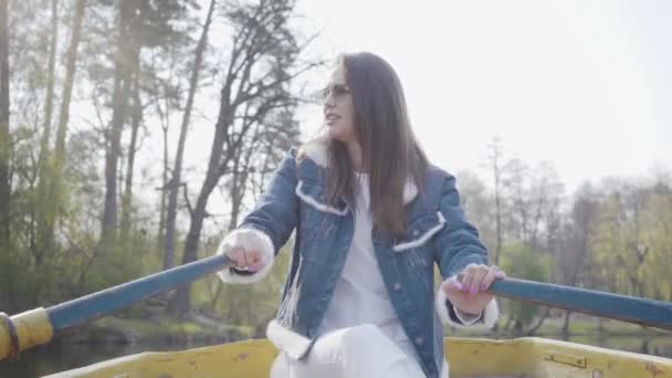 Portrait of a charming girl in glasses and a denim jacket floating on a boat on a lake or river. Beautiful brunette is actively relaxing on a day off or traveling enjoying nature. — Stock Video