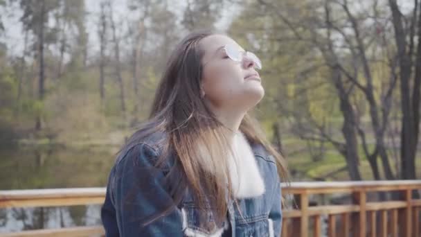 Portrét rozkošné mladé ženy v džínách, které stály na dřevěném mostě a rozhlédne se po kameře a pak sunou sluneční brýle. Dívka obdivující krásnou krajinu. Volný čas venku. — Stock video