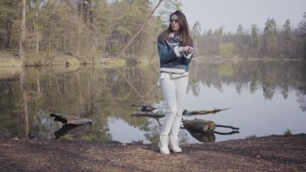 川岸に立つ白いズボン、ジーンズジャケット、サングラスを着たグラマー女性。その少女は冷たく、自分を暖めようと手をこすりつける。背景に美しい風景 — ストック動画