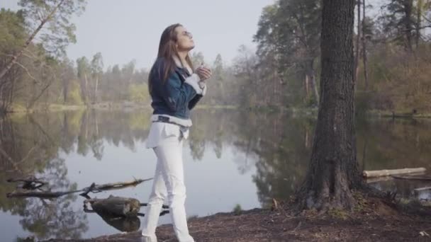 Bella giovane donna in pantaloni bianchi, giacca jeans e occhiali da sole in piedi sulla riva del fiume. La ragazza ha freddo, si strofina le mani cercando di riscaldarsi. Bellissimo paesaggio su sfondo — Video Stock