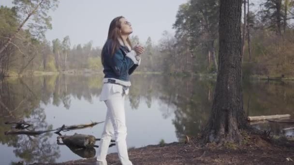 Mujer joven y bonita en pantalones blancos, chaqueta vaquera y gafas de sol de pie en la orilla del río. La chica tiene frío, se frota las manos tratando de calentarse. Hermoso paisaje en el fondo — Vídeos de Stock