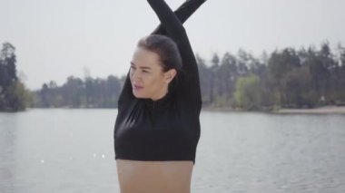 Çekici ince kız spor egzersizleri yapıyor, nehir kıyısında duruyor. Arka planda güzel manzara. Boş zaman, aktif yaşam tarzı, egzersiz. Kamera aşağı hareket ediyor