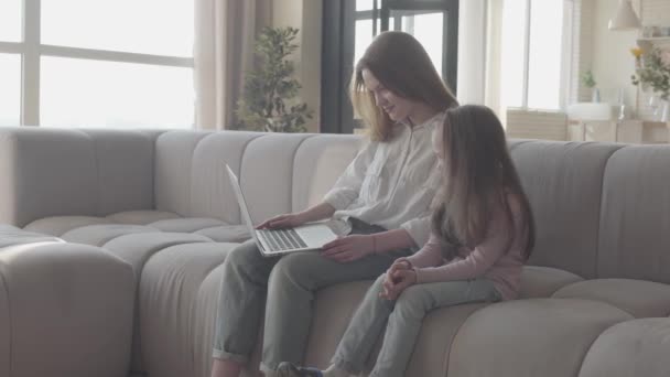 Uma jovem mãe e filha com cabelos longos sentados juntos no sofá na sala de estar digitando em um laptop. A criança está perto da mãe, ajudando-a a digitar. Lazer de mulher e seu filho . — Vídeo de Stock