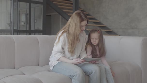 Gyönyörű fiatal anya és az ő aranyos kis lánya használ egy tablettát, és mosolyogva, ül a kanapén a nagy házban. Családi kapcsolatok. — Stock videók