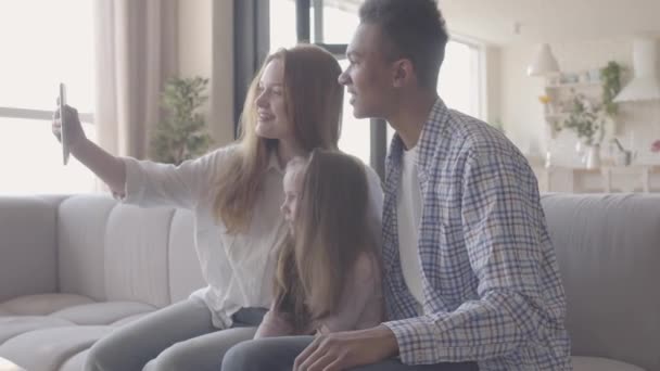 Joven mujer rubia caucásica sentarse con el hombre negro africano y entre ellos es pequeña hija adorable con el pelo largo y claro. Familia internacional haciendo una foto por tableta. Relaciones familiares . — Vídeo de stock