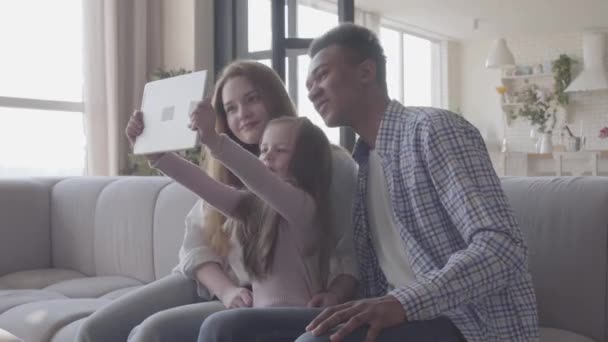 Belle jeune famille internationale à la maison, homme afro-américain, femme caucasienne et petite fille assise sur le canapé avec tablette. L'enfant prend selfie en famille en utilisant un comprimé. Famille — Video