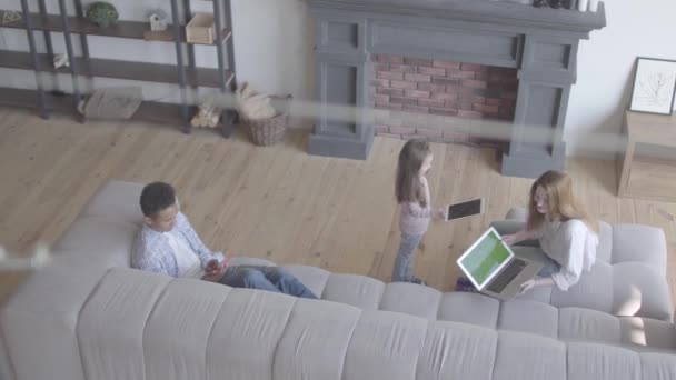 Beautiful young international family at home, african american man, caucasian woman and girl in the large living room using gadgets. A girl comes to mum giving her tablet. Top view — Wideo stockowe