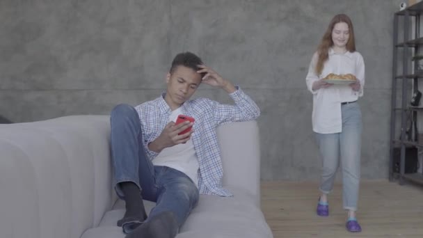 Young african american man sitting on the sofa, caucasian woman bringing him the croissants on the plate. The man typing on cellphone. Happy family resting at home. Multicultural couple — 비디오