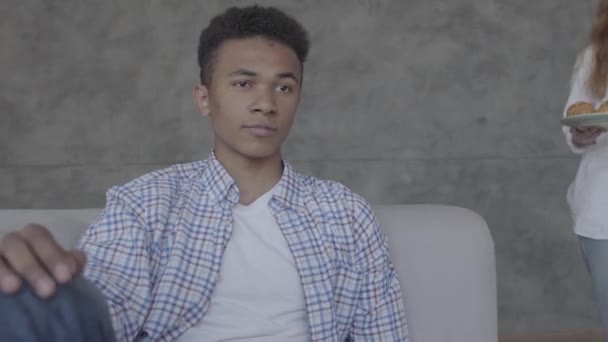 Portrait de jeune homme afro-américain assis sur le canapé, femme caucasienne lui apportant les croissants sur l'assiette gros plan. Bonne famille reposant à la maison. Couple multiculturel — Video