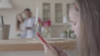 Küçük kız cep telefonu ile oynamak ve arka planda büyük mutfakta annesi ve babası