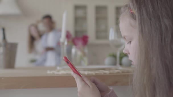 Μικρό κορίτσι παίζουν με το κινητό τηλέφωνο και στο παρασκήνιο είναι η μητέρα και ο πατέρας της στη μεγάλη κουζίνα — Αρχείο Βίντεο