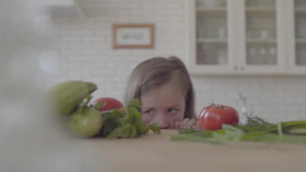 Belle petite fille regardant la table avec des tomates, des courgettes, des légumes verts, des oignons. Un gamin qui prend une tomate et court. — Video