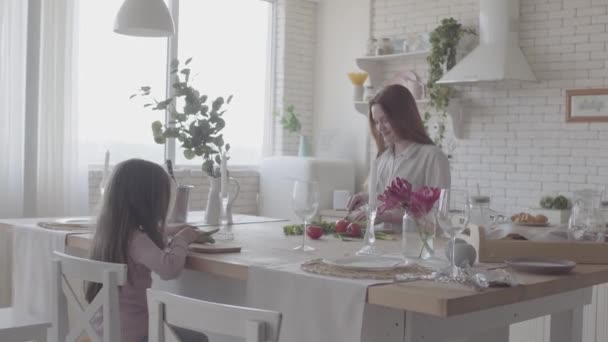 Mulher sorridente bonita cozinhando salada de pé à mesa na cozinha moderna. Menina sentada na cadeira ajudando sua mãe, cortando abobrinha. Família simpática e feliz. Relações familiares — Vídeo de Stock