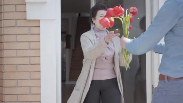 Volwassen kleinzoon een bezoek aan zijn grootmoeder, het brengen van haar boeket van tulpen. De bebaarde man knuffelen zijn oma. Mensen glimlachen. Vrouw uitnodigt vent in huis. Familie relatie, verjaardag vieren — Stockvideo