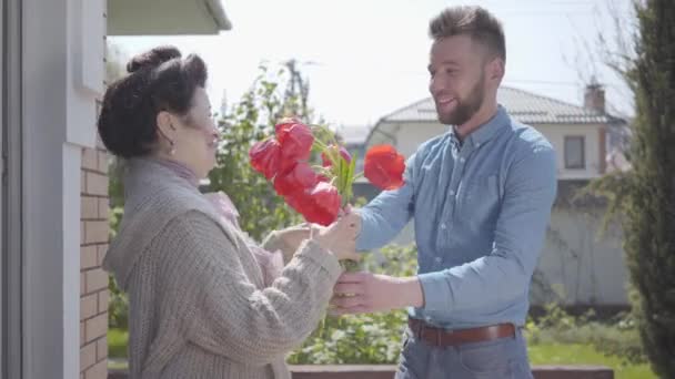 Дорослий онук відвідує бабусю, приносить свій букет тюльпанів. Бородатий чоловік обіймає бабусю. Люди посміхаються. Жінка запрошує хлопця всередину будинку. Сімейні стосунки, святкування дня народження — стокове відео