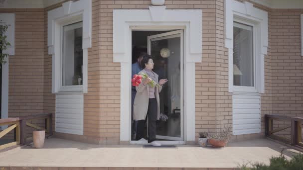 Dospělý vnuk a jeho babička, kteří drží kytici tulipány, shlížejí na verandu velkého domu. Bábi vypráví příběh muži, oba se usmívající. Rodinný vztah — Stock video