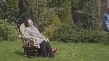 Portre sevimli olgun bayan güneş zevk sallanan sandalyede çim üzerinde oturan. Yetişkin torun sulama olabilir, kadın çalıları sulamasını istiyor. Bir ile büyükanne yardım adam