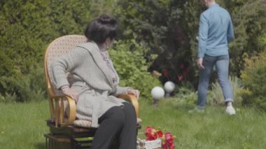 Portre sevimli olgun bayan güneş zevk sallanan sandalyede çim üzerinde oturan. Çimenlerin üzerindeki kadının yanında lalelerin olduğu sepet. Bahar. Büyükanne dışarıda dinleniyor
