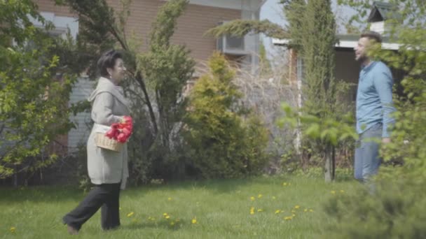 Взрослая женщина идет с корзиной с тюльпанами на заднем дворе. Приходит взрослый внук и обнимает даму, парня, навещающего свою бабушку. Семейное собрание, дружеские отношения — стоковое видео