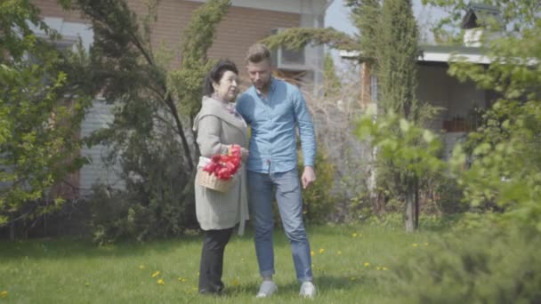 Retrato mujer madura con la cesta con tulipanes en las manos hablando con su nieto en el patio trasero. Nieto adulto y la abuela discutiendo el jardín, luego se van. Encuentro familiar, amistoso — Vídeo de stock