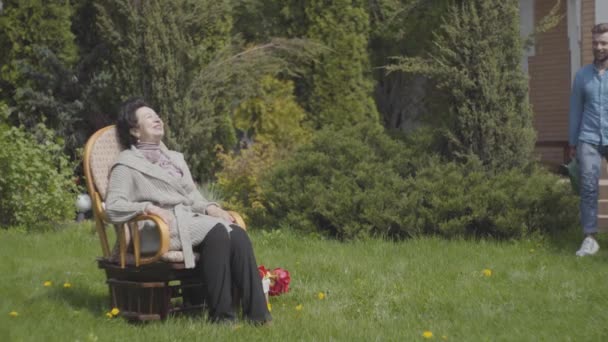 Portrait dame mature assise sur la pelouse dans le fauteuil à bascule profitant du soleil. Petit-fils adulte venant avec l'arrosoir, la femme lui demandant d'arroser le buisson. L'homme aidant mamie avec un jardin — Video