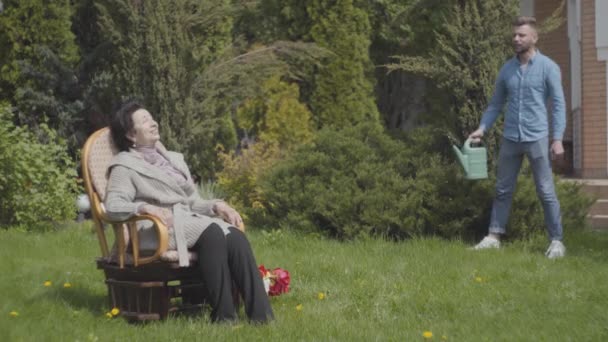 Retrato mujer adulta sentada en el césped en la mecedora disfrutando del sol. El nieto adulto viene con la regadera, la mujer le pide que riegue el arbusto. El hombre ayudando a la abuela con un jardín — Vídeos de Stock