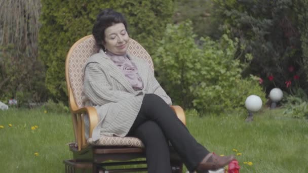 Portrétní dospělá žena, která sedí na trávníku v houpacím křesle a užívá si slunce zblízka. Dospělý vnuk zakrývající oči ženy, usmívající se a objetí. Muž, který navštívil svou babičku — Stock video