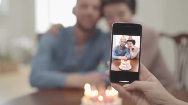 Retrato de la mano femenina haciendo una foto en el celular de la feliz sonrisa de la mujer madura abrazándose con el nieto adulto, ambos en gorras de cumpleaños. El pastel está sobre la mesa delante de la gente. Cumpleaños de — Vídeo de stock