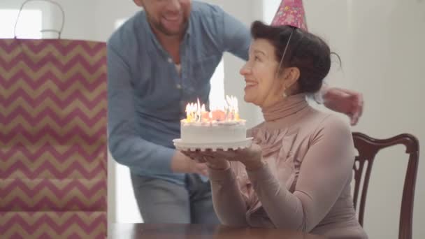 Porträt einer emotional lächelnden Dame, die am Tisch sitzt und die kleine Torte mit den vielen Kerzen hält. Der erwachsene Enkel umarmt die Frau und bläst gemeinsam die Kerzen aus. Geburtstag der Seniorin — Stockvideo