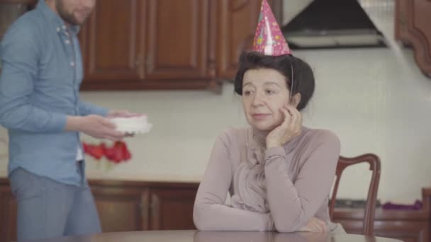 그녀의 머리에 생일 모자와 테이블에 앉아 성숙한 여자. 성인 손자는 작은 케이크를 가져 와서 여자 앞에서 테이블에 놓고 할머니를 껴안고 행복하고 미소지습니다. — 비디오
