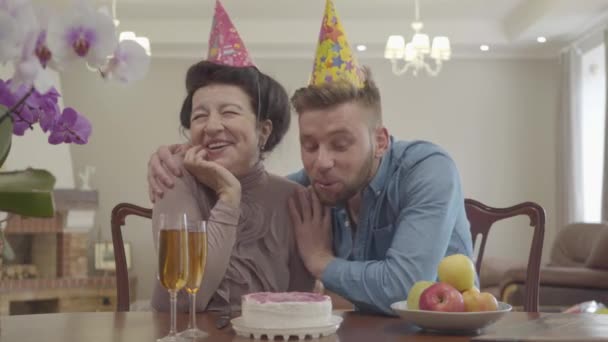 Portret dojrzała kobieta i dorosły wnuk rozmawiając siedząc przy stole z urodzinowym kapelusza na głowach. Na stole znajdują się małe ciasto, kieliszki do soku, talerz z jabłkami. Uroczystość urodzinowa — Wideo stockowe