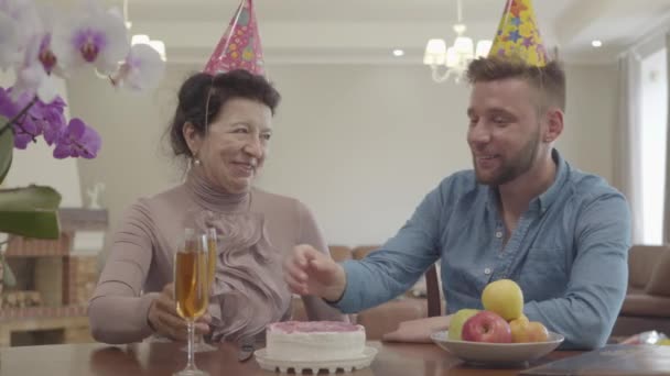 Retrato abuelita y nieto adulto bebiendo jugo sentado en la mesa con gorra de cumpleaños en la cabeza. En la mesa, hay pastel y plato con manzanas. Celebración de cumpleaños. Relación familiar — Vídeo de stock