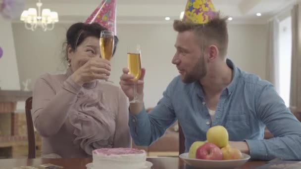 Linda abuela y nieto adulto tintineo los vasos sentados en la mesa con gorra de cumpleaños en sus cabezas. Mujer madura y hombre más joven bebiendo jugo. Celebración de cumpleaños. Relación familiar — Vídeo de stock