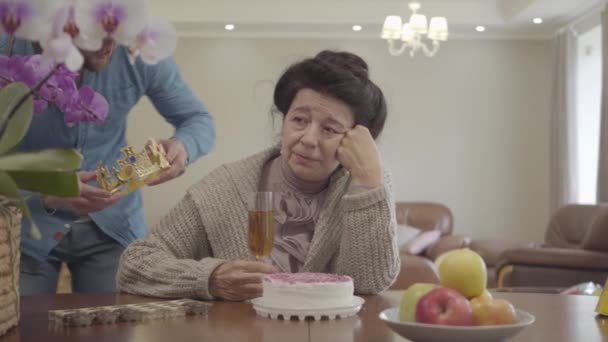 Mature lady sitting at the table in front of small cake and apples on the plate. Adult grandson brings the crown and puts it on the head of woman, then hugging the granny, both happy and smiling — Stock Video